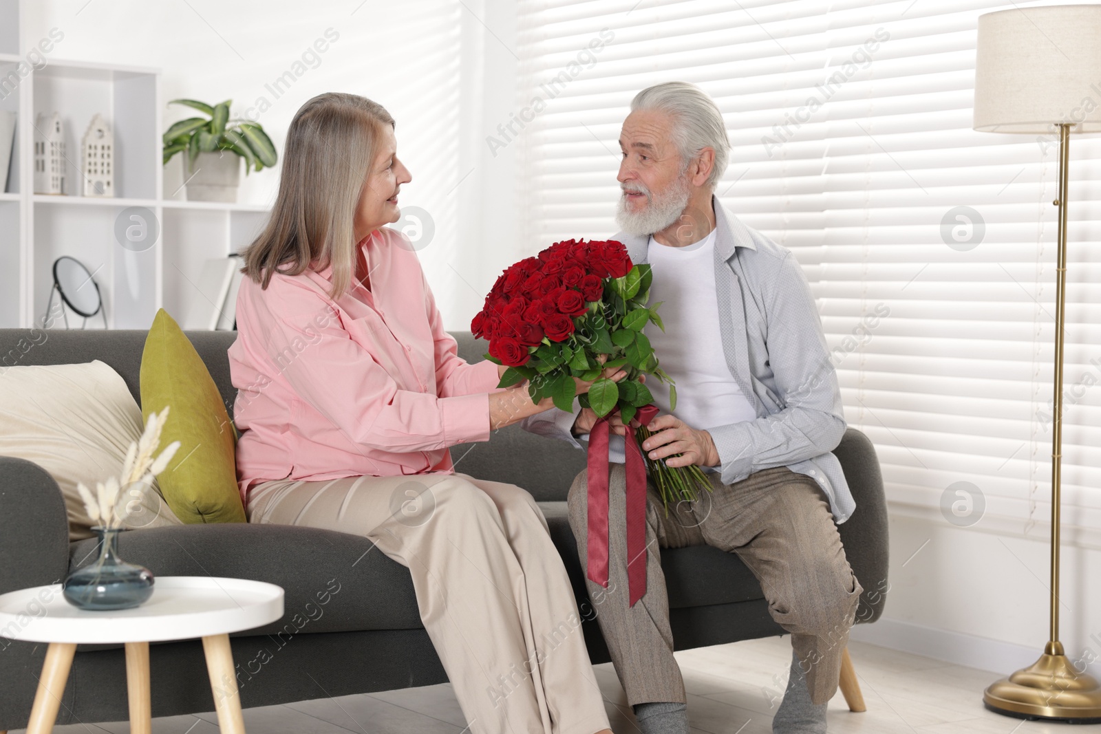 Photo of Man presenting bouquet of roses to his happy wife on sofa at home