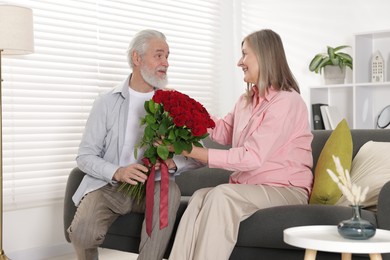 Photo of Man presenting bouquet of roses to his happy wife on sofa at home