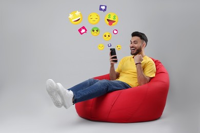 Image of Social media. Happy man sitting in beanbag and using mobile phone on light grey background. Different notification signs and emojis near gadget