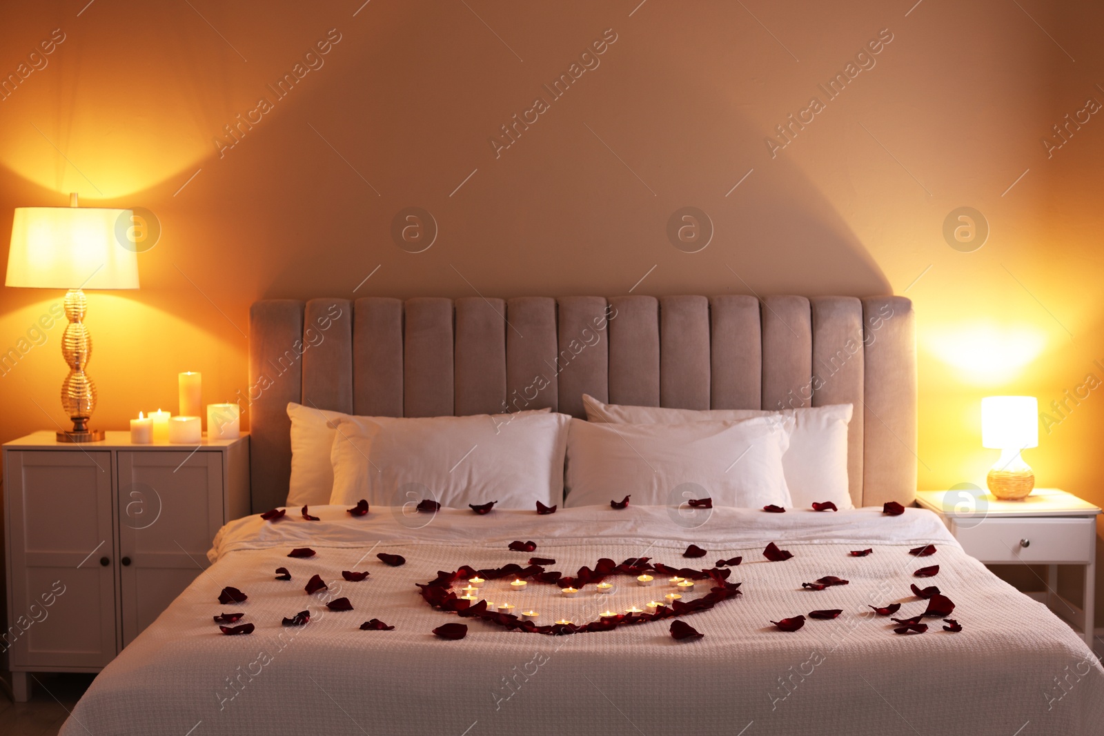 Heart made with beautiful rose petals and burning candles on bed indoors Photo of Heart made with beautiful rose petals and burning candles on bed indoors