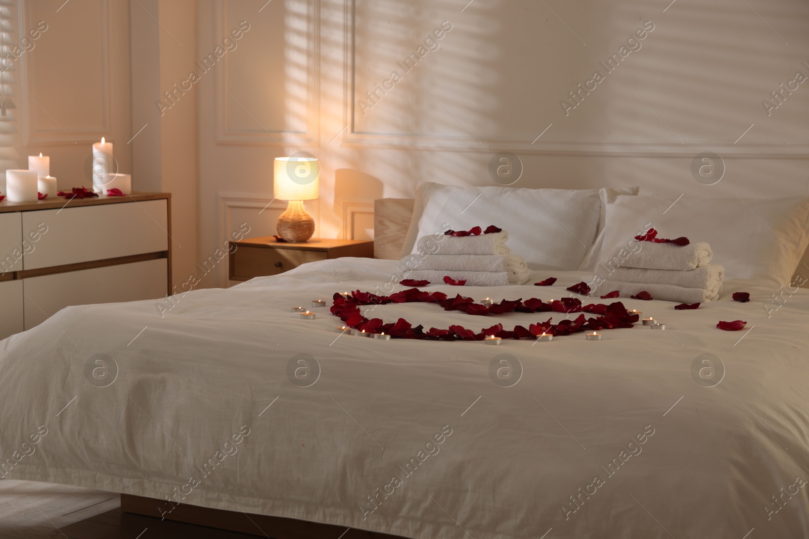 Heart made with rose petals and burning candles on bed indoors Photo of Heart made with rose petals and burning candles on bed indoors