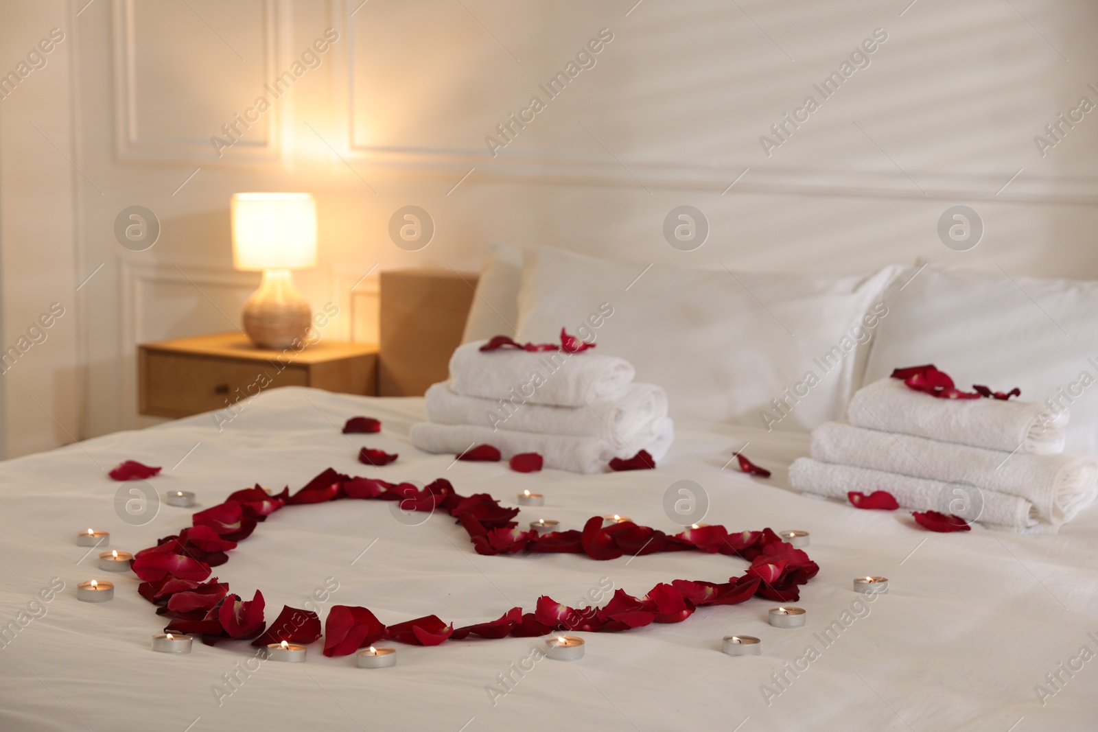 Heart made with rose petals and burning candles on bed indoors Photo of Heart made with rose petals and burning candles on bed indoors