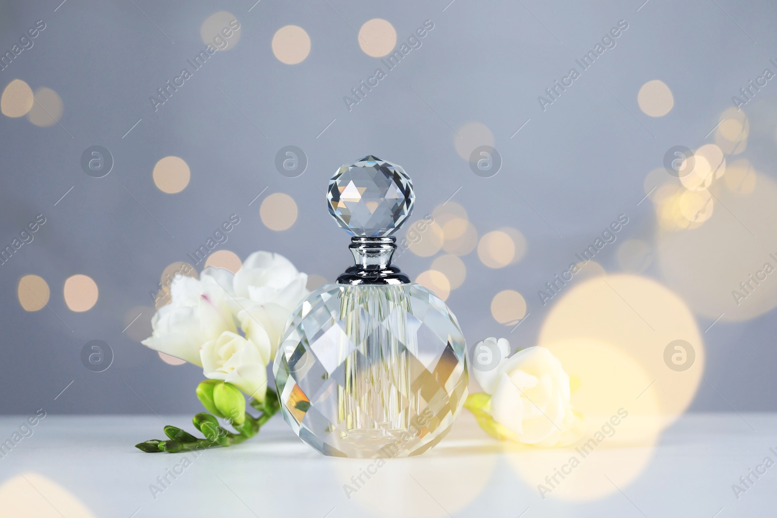 Bottle of aromatic perfume and flowers on light table against grey background with blurred lights, closeup. Bokeh effect Photo of Bottle of aromatic perfume and flowers on light table against grey background with blurred lights, closeup. Bokeh effect
