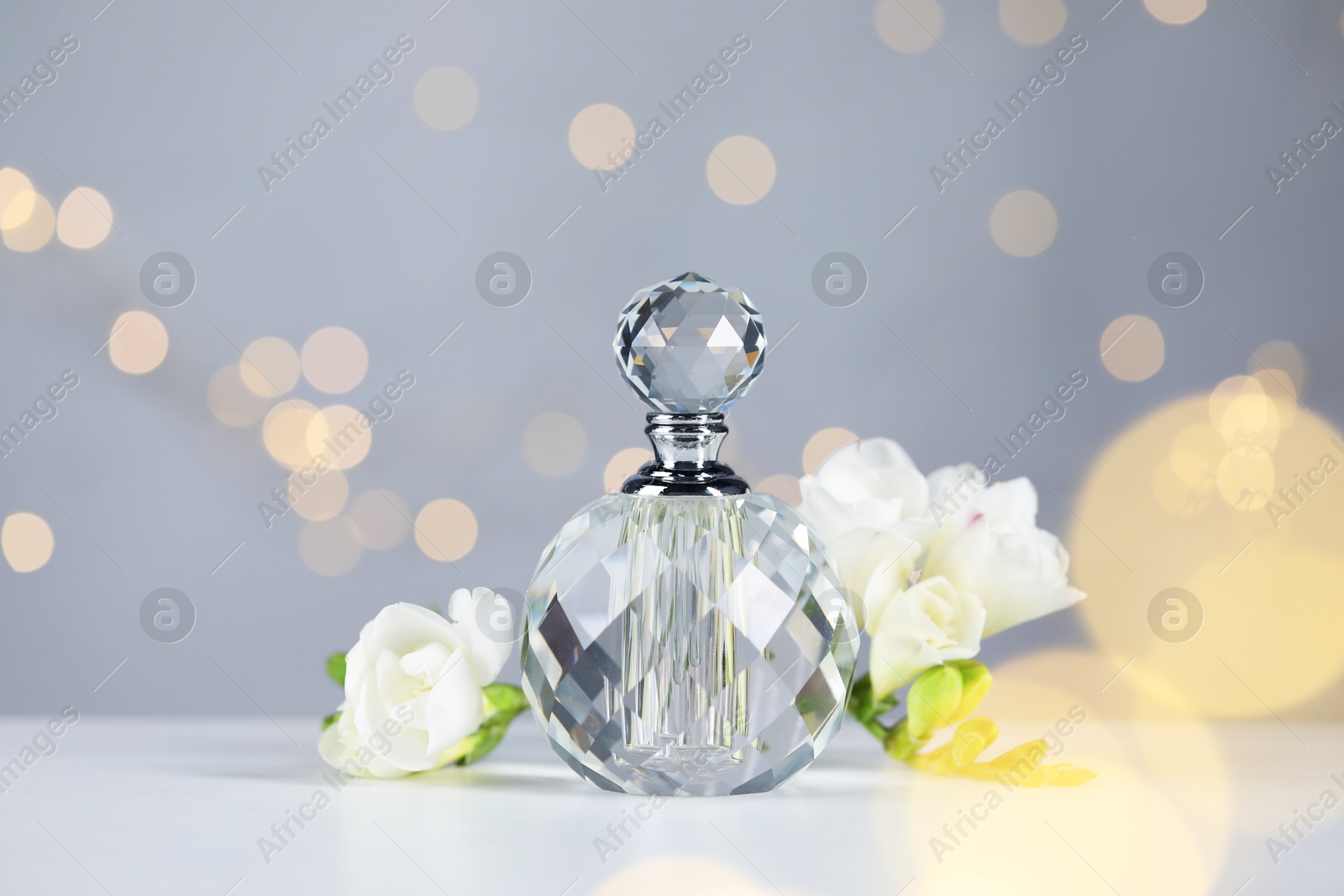 Bottle of aromatic perfume and flowers on light table against grey background with blurred lights, closeup. Bokeh effect Photo of Bottle of aromatic perfume and flowers on light table against grey background with blurred lights, closeup. Bokeh effect