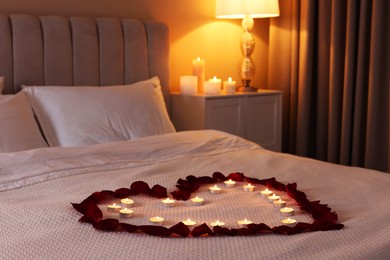 Photo of Heart made with beautiful rose petals and burning candles on bed indoors