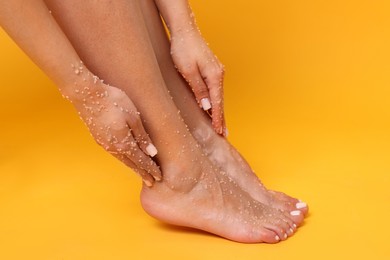 Photo of Woman applying body scrub onto her feet on orange background, closeup