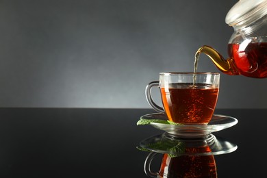 Photo of Pouring freshly brewed tea from teapot into cup at dark mirror table, closeup. Space for text
