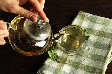 Photo of Woman pouring freshly brewed tea from teapot into cup at wooden table, closeup
