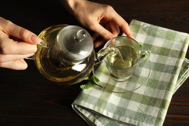 Photo of Woman pouring freshly brewed tea from teapot into cup at wooden table, closeup