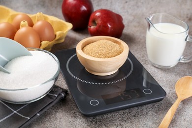 Photo of Digital kitchen scale with bowl of brown sugar and other products on grey table, closeup