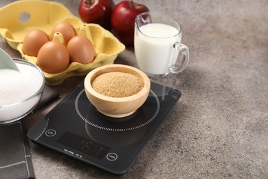 Photo of Digital kitchen scale with bowl of brown sugar and other products on grey table, closeup. Space for text