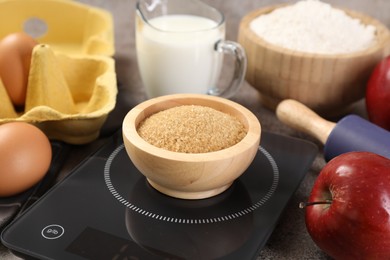 Photo of Digital kitchen scale with bowl of brown sugar and other products on grey table, closeup