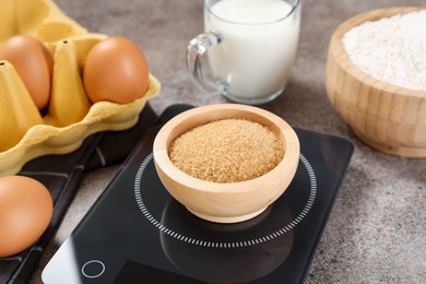 Photo of Digital kitchen scale with bowl of brown sugar and other products on grey table, closeup