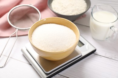 Photo of Digital kitchen scale with bowl of sugar and other products on white wooden table, closeup