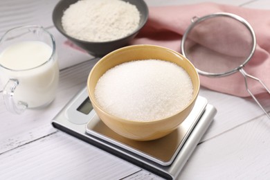 Photo of Digital kitchen scale with bowl of sugar and other products on white wooden table, closeup