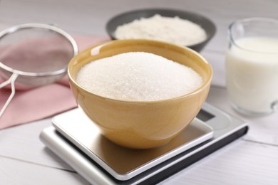 Photo of Digital kitchen scale with bowl of sugar and other products on white wooden table, closeup