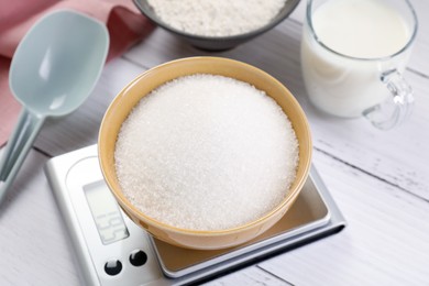 Photo of Digital kitchen scale with bowl of sugar and other products on white wooden table, closeup