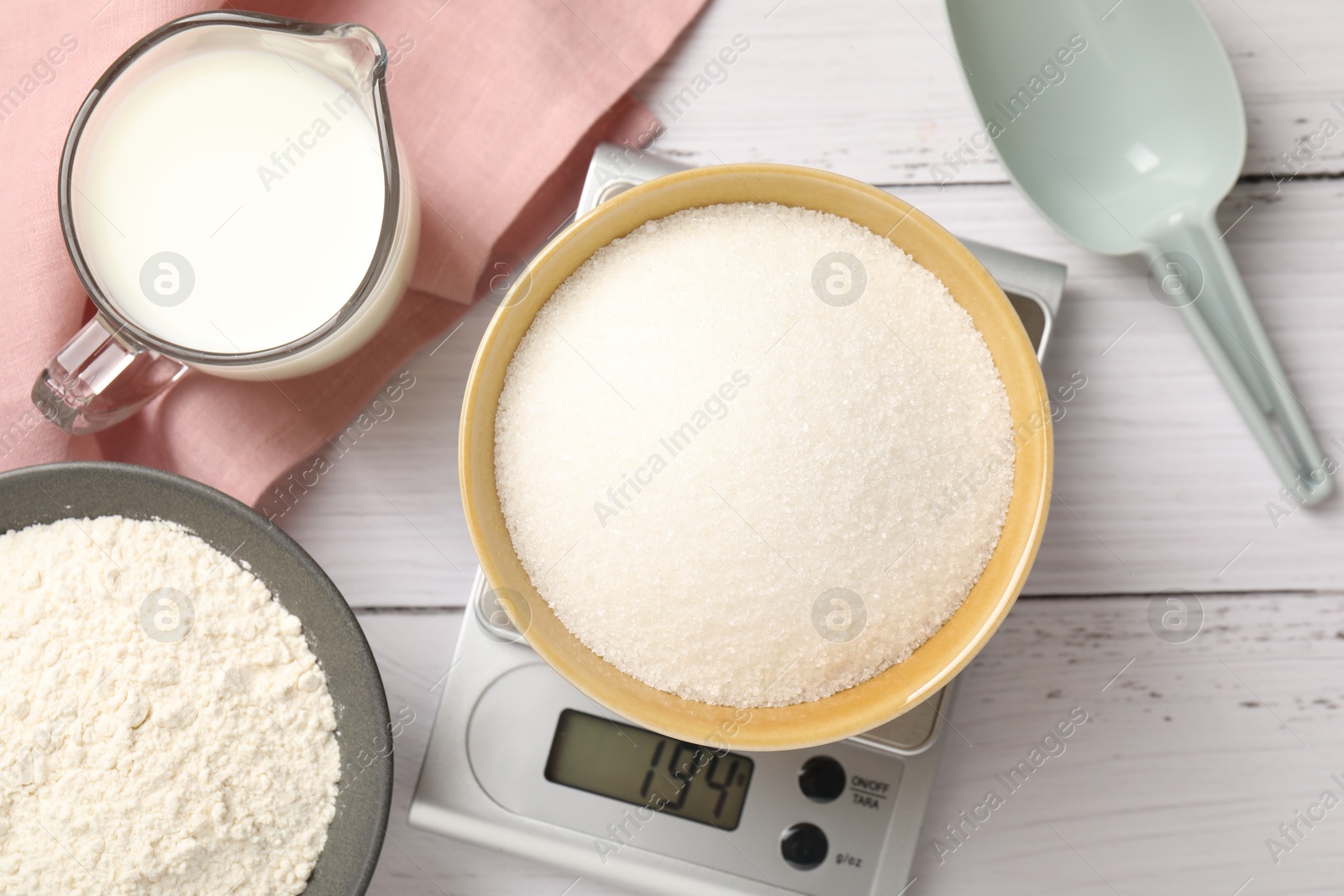 Digital kitchen scale with bowl of sugar and other products on white wooden table, flat lay Photo of Digital kitchen scale with bowl of sugar and other products on white wooden table, flat lay