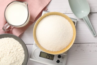 Photo of Digital kitchen scale with bowl of sugar and other products on white wooden table, flat lay