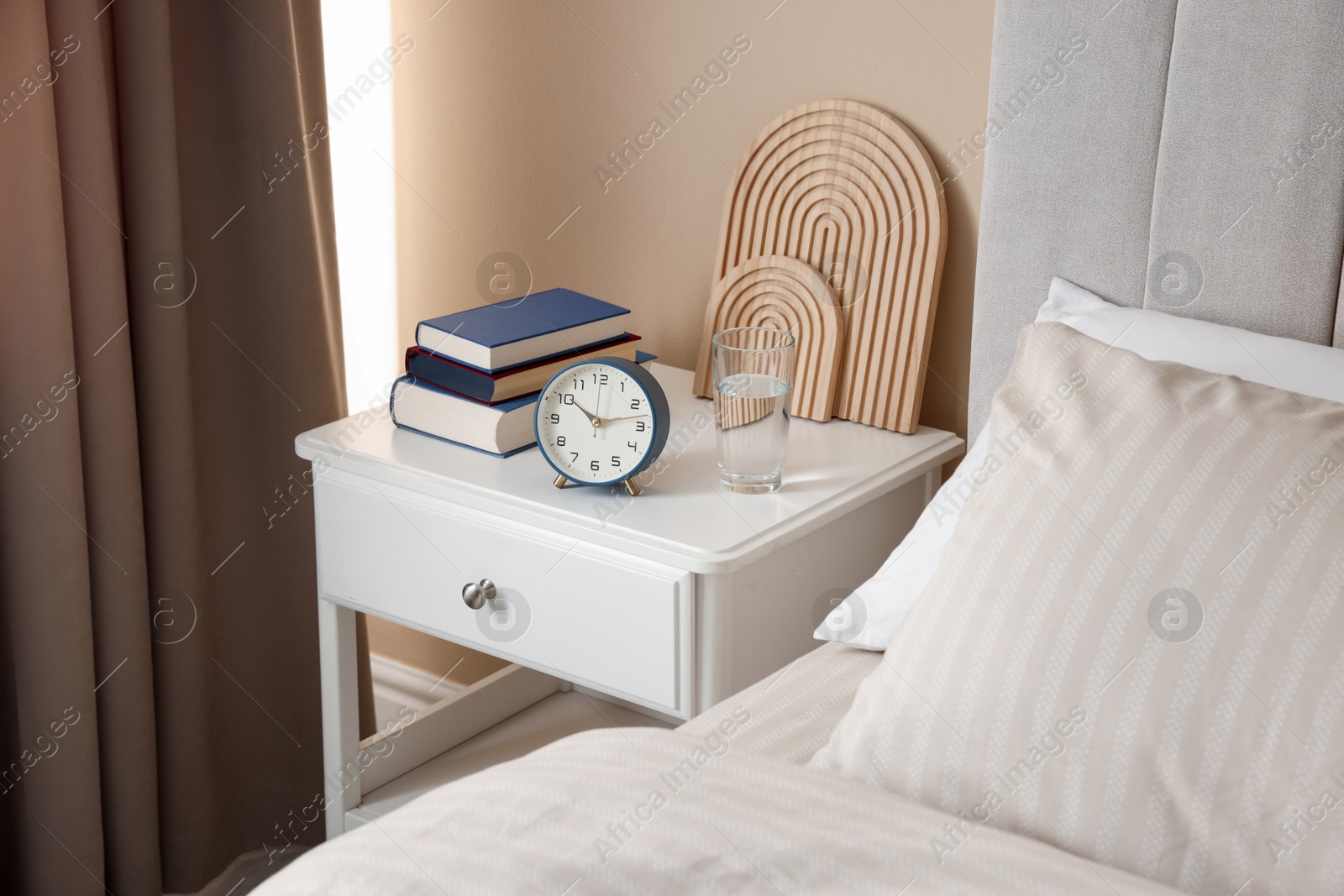 Bedside table with books, decor, glass of water and alarm clock near bed in room Photo of Bedside table with books, decor, glass of water and alarm clock near bed in room