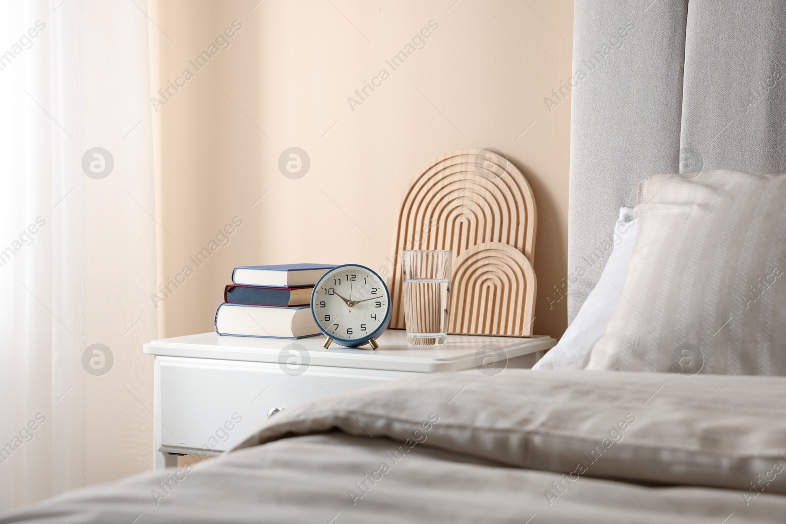 Bedside table with books, decor, glass of water and alarm clock near bed in room Photo of Bedside table with books, decor, glass of water and alarm clock near bed in room