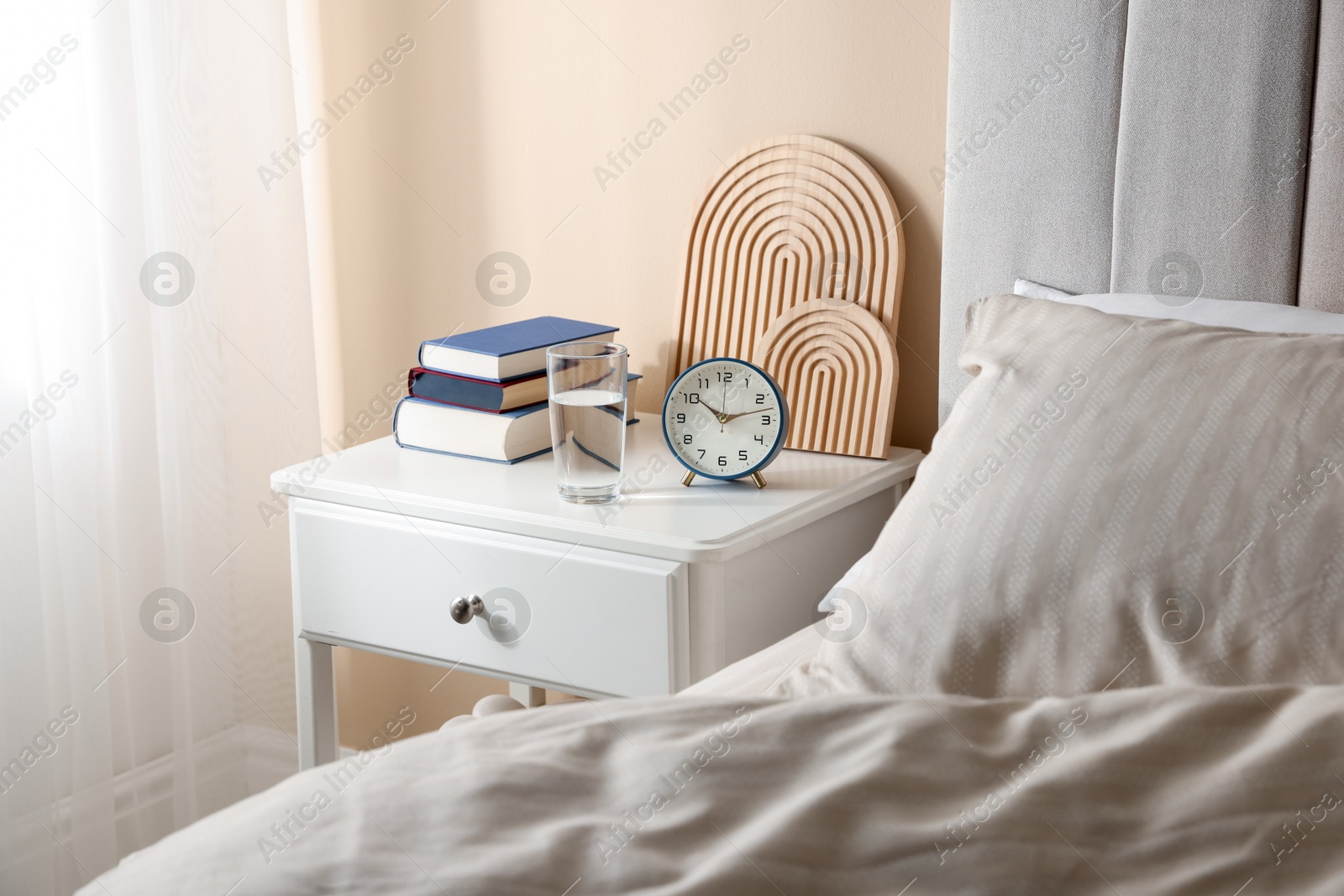 Bedside table with books, decor, glass of water and alarm clock near bed in room Photo of Bedside table with books, decor, glass of water and alarm clock near bed in room