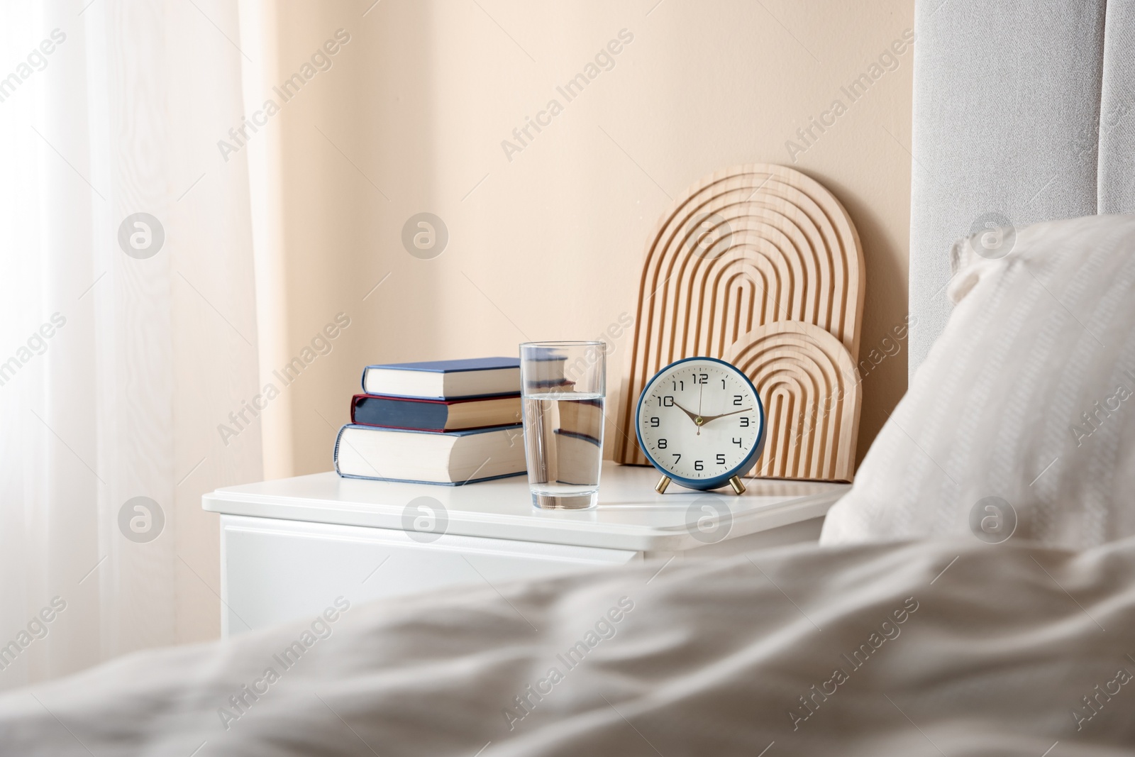 Bedside table with books, decor, glass of water and alarm clock near bed in room Photo of Bedside table with books, decor, glass of water and alarm clock near bed in room