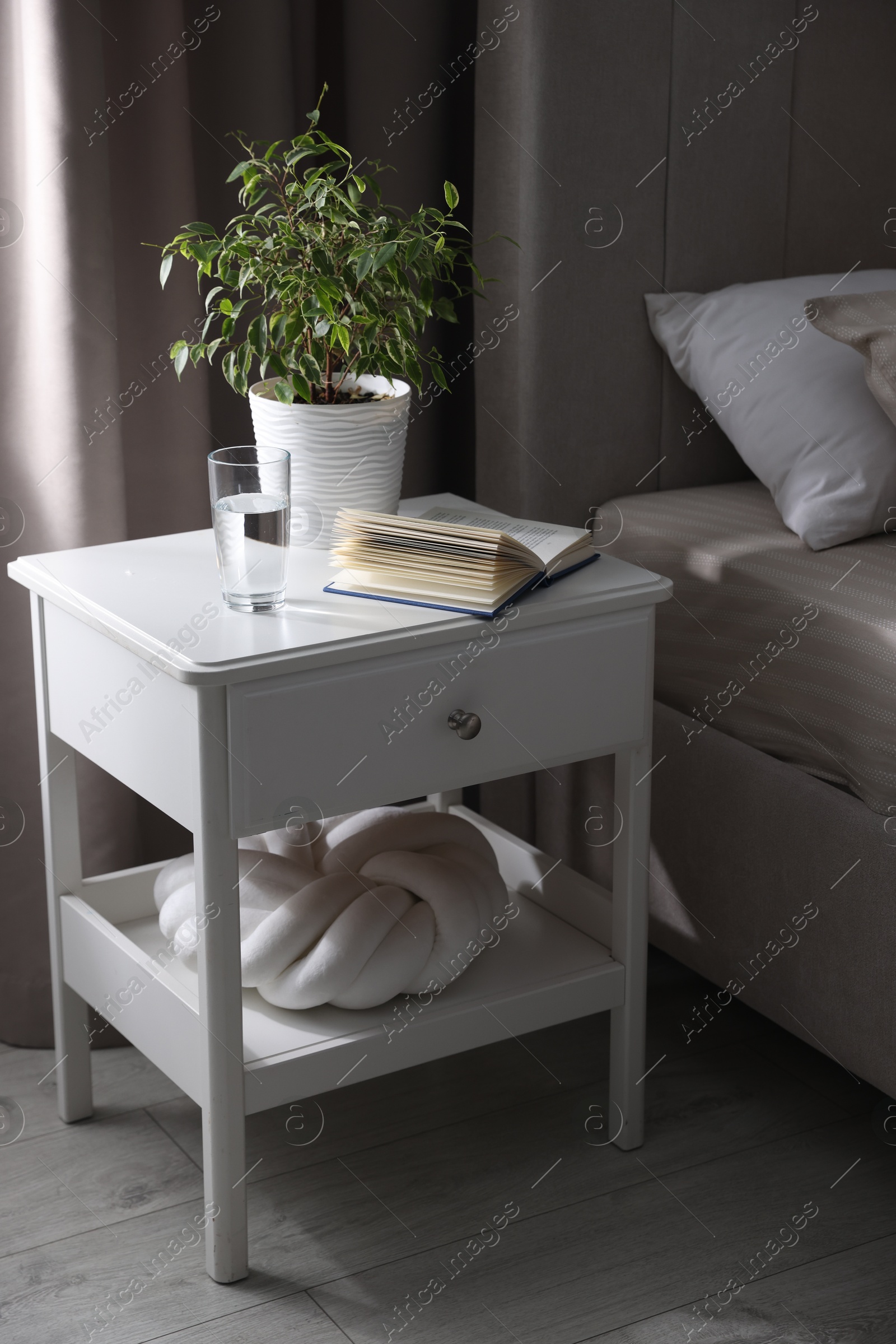 Bedside table with book, houseplant and glass of water near bed in room Photo of Bedside table with book, houseplant and glass of water near bed in room