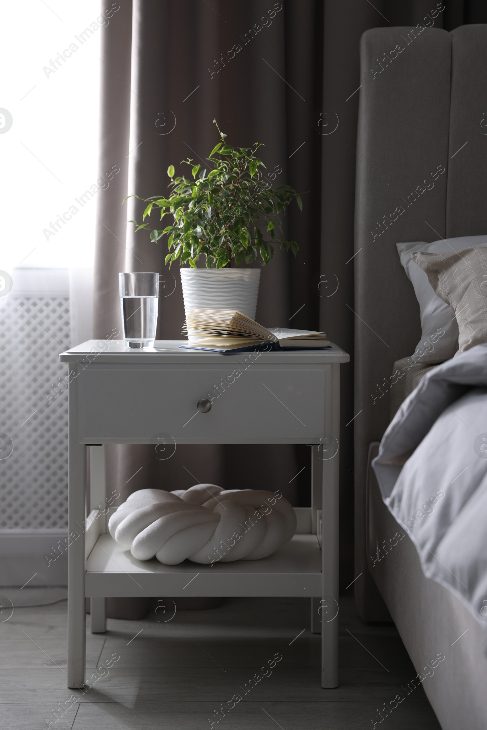 Bedside table with book, houseplant and glass of water near bed in room Photo of Bedside table with book, houseplant and glass of water near bed in room