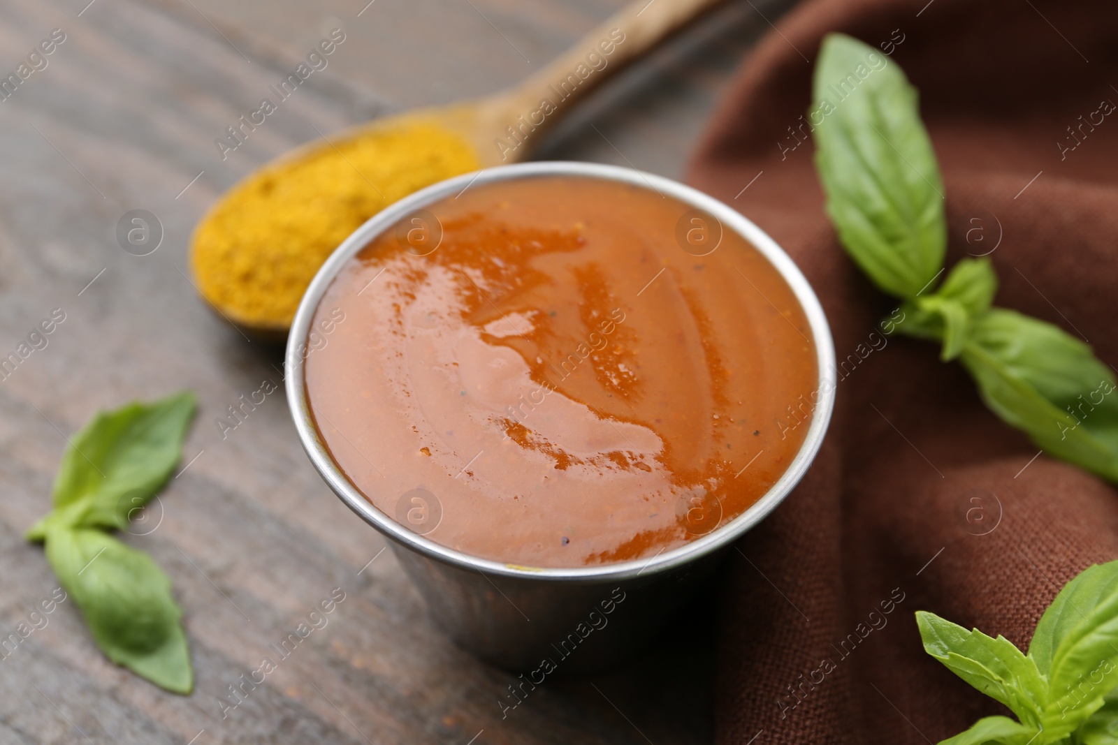 Photo of Tasty curry sauce in metal dish, powder and basil on wooden table, closeup