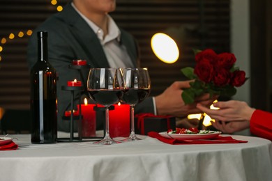 Photo of Man giving roses to his girlfriend in restaurant, focus on glasses of red wine. Romantic dinner