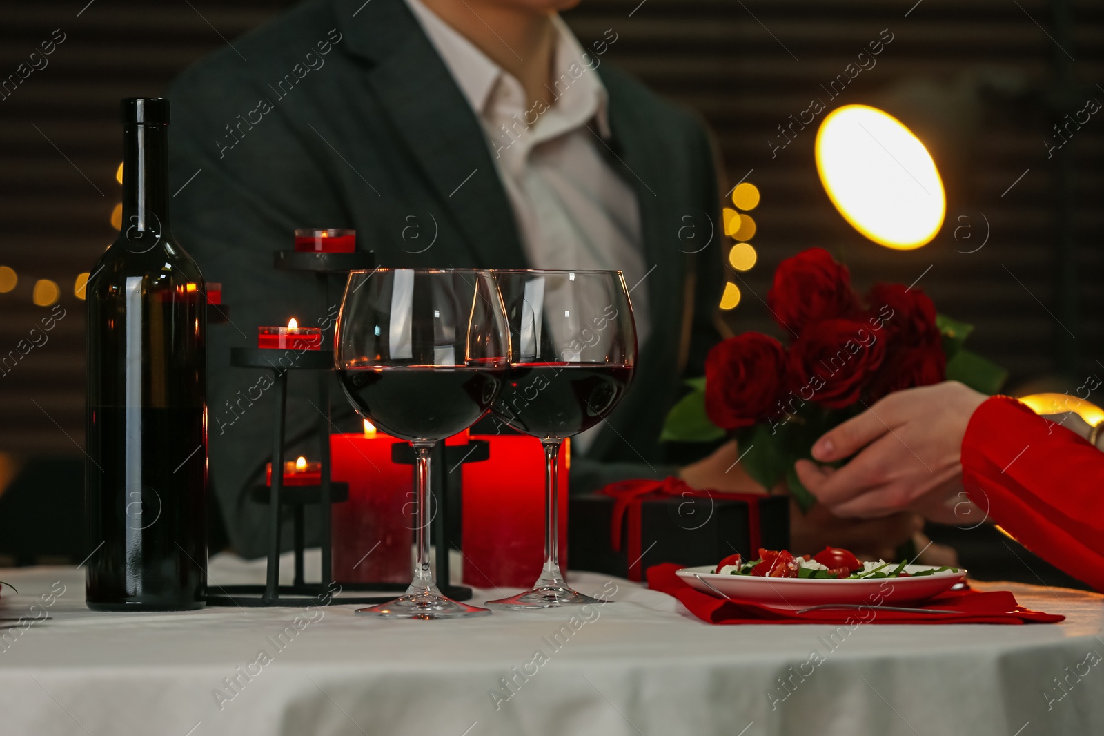 Photo of Man giving roses to his girlfriend in restaurant, focus on bottle and glasses of red wine. Romantic dinner