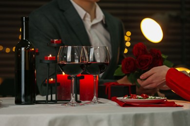 Photo of Man giving roses to his girlfriend in restaurant, focus on bottle and glasses of red wine. Romantic dinner