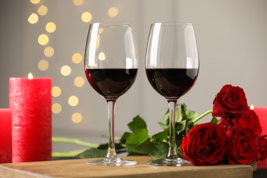 Photo of Glasses of red wine, burning candle and roses on table against gray background with blurred lights. Romantic dinner