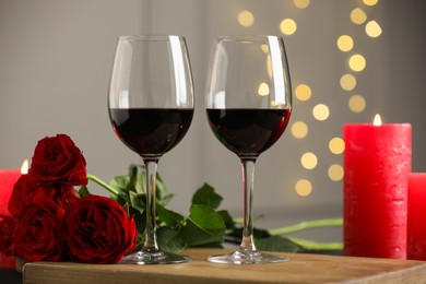 Photo of Glasses of red wine, burning candle and roses on table against gray background with blurred lights. Romantic dinner