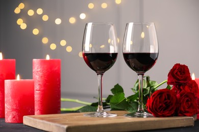 Photo of Glasses of red wine, burning candles and roses on table against gray background with blurred lights. Romantic dinner