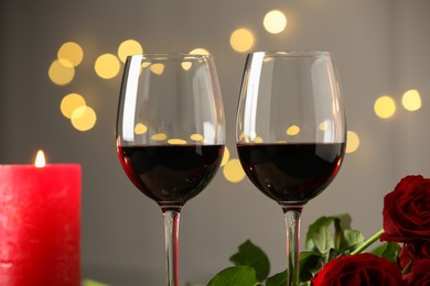 Photo of Glasses of red wine, burning candle and roses against blurred background, closeup. Romantic dinner