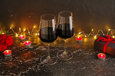 Photo of Glasses of wine, burning candles, gift box and roses on black textured table. Romantic dinner