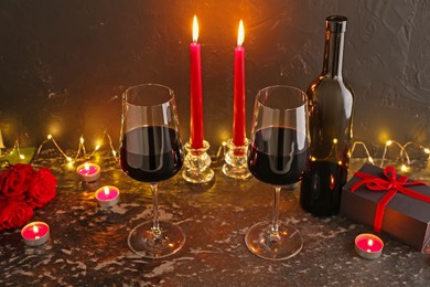 Photo of Glasses of wine, burning candles, gift box and roses on black textured table. Romantic dinner