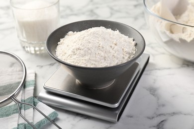 Photo of Digital kitchen scale with bowl of flour and other products on white marble table, closeup