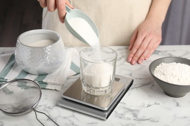 Photo of Woman putting sugar into glass on digital kitchen scale at white marble table, closeup