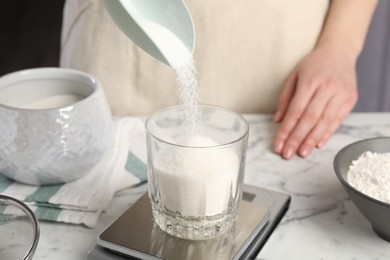 Photo of Woman putting sugar into glass on digital kitchen scale at white marble table, closeup