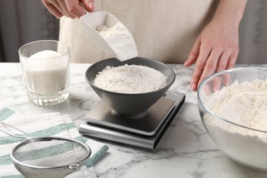 Photo of Woman putting sugar into bowl on digital kitchen scale at white marble table, closeup