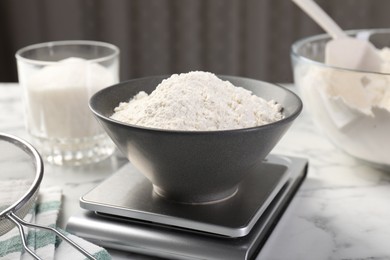 Photo of Digital kitchen scale with bowl of flour and other products on white marble table, closeup