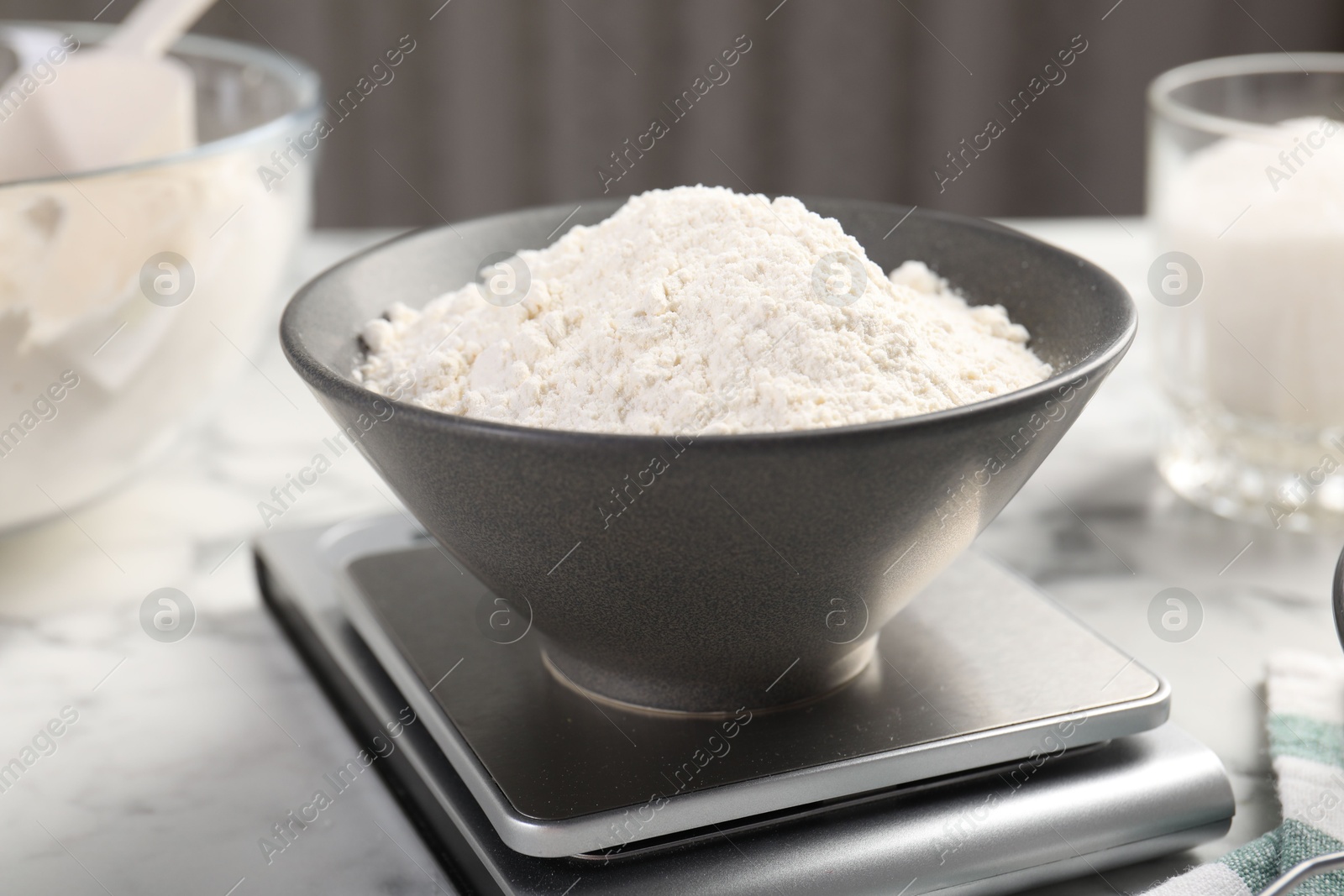 Photo of Digital kitchen scale with bowl of flour and other products on white marble table, closeup
