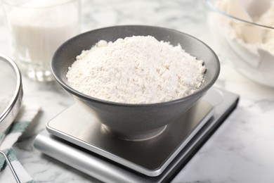 Photo of Digital kitchen scale with bowl of flour and other products on white marble table, closeup