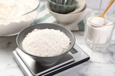Photo of Digital kitchen scale with bowl of flour and other products on white marble table, closeup