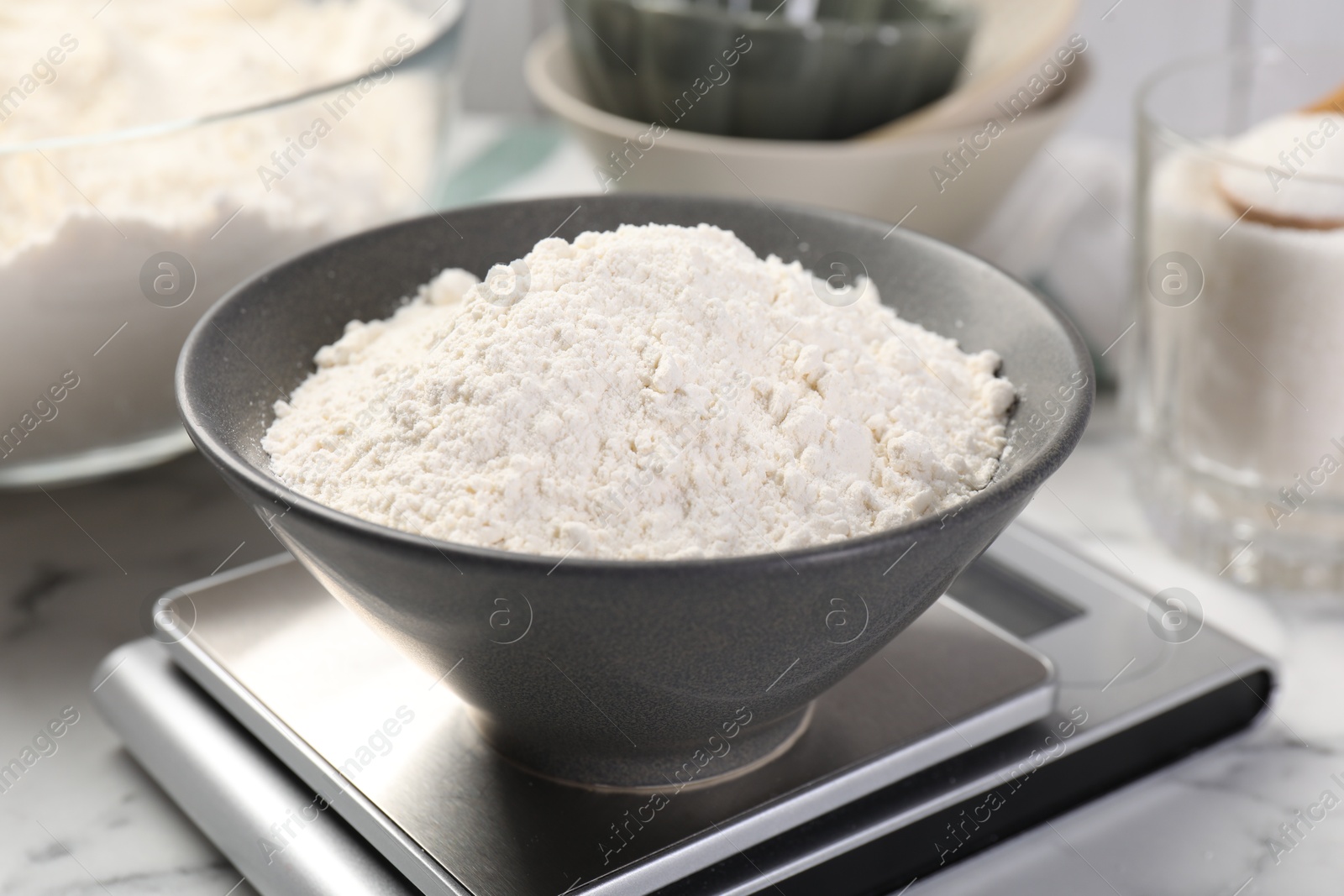 Photo of Digital kitchen scale with bowl of flour and other products on white marble table, closeup