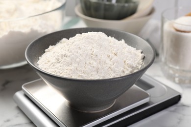 Photo of Digital kitchen scale with bowl of flour and other products on white marble table, closeup