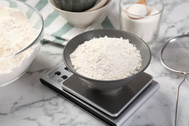 Photo of Digital kitchen scale with bowl of flour and other products on white marble table, closeup